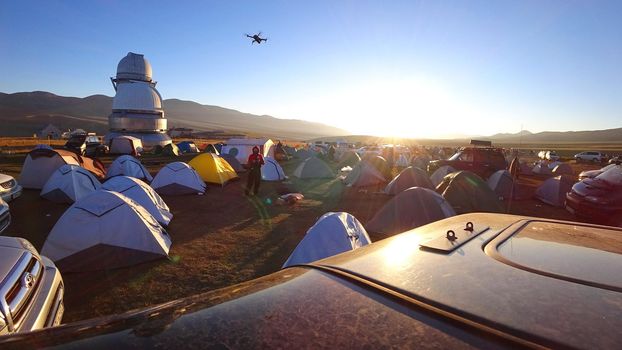 Sunrise over the camp near the Assi Observatory. The sun comes out from behind the high hills. Red-yellow color. Lots of tents and cars. The guy launches the drone. Telescopes are covered with domes
