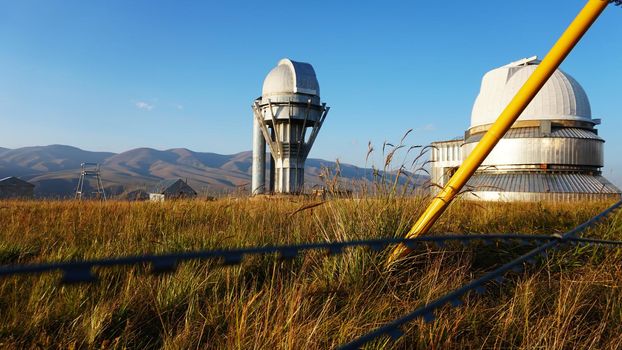 A large observatory is protected by a barbed fence. There is a long sharp wire around perimeter. Buildings with domes and a telescope are visible in distance. Yellow-green grass. Mountainous terrain