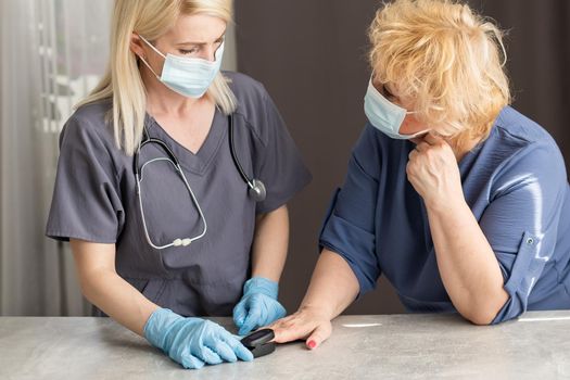 Medical help at home. Woman doctor in a medical mask measures the patient's pulse and oxygen saturation to middle aged woman using a pulse oximeter while sitting on sofa, healthcare concept.