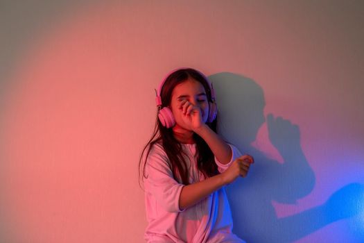 A dancing little girl in pink headphones with loose dark hair, in neon pink blue light near the wall