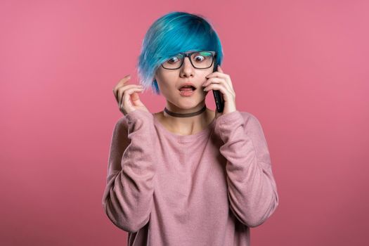 Unusual woman with blue hair making call with smartphone over pink wall background. Girl nervous, expresses shock and fear about bad news.