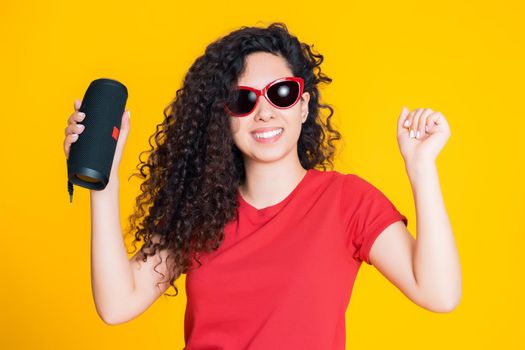 Young beautiful woman with curly hairstyle enjoying and dancing at yellow background. Modern trendy girl listening to music by wireless portable speaker