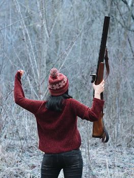 Hunter woman in burgundy warm clothes with gun. Girl hunting in the forest and rejoices her victory by raising rifle up after firing. High quality photo