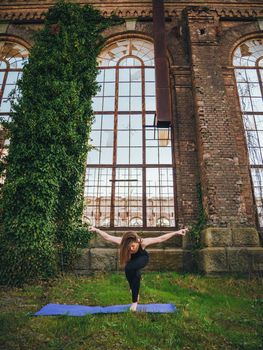 Young girl in black bodysuit is practicing yoga near an old abandoned building with large windows without glass and overgrown with green ivy. Healthy lifestyle concept. High quality photo