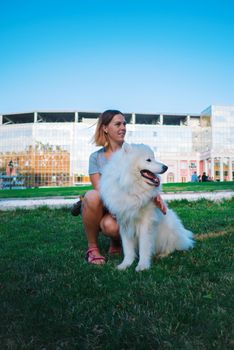 An adult woman with red hair plays and strokes her dog of the Samoyed breed. White fluffy pet in a park with mistress on a green lawn have fun. High quality photo