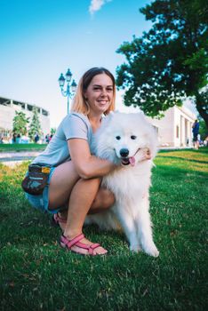 An adult woman with red hair plays and strokes her dog of the Samoyed breed. White fluffy pet in a park with mistress on a green lawn have fun. High quality photo