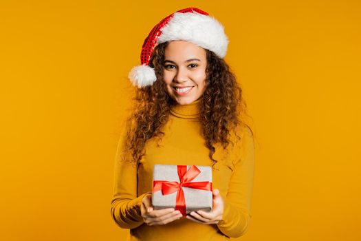 Excited woman in Santa hat received gift box with bow. She is happy and flattered by attention. Girl with present on yellow background. Studio shot. High quality photo