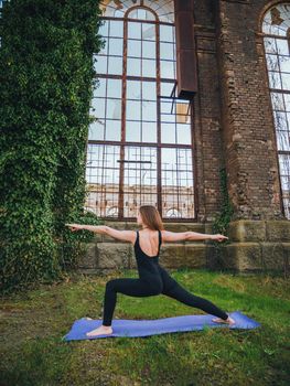 Young girl in black bodysuit is practicing yoga near an old abandoned building with large windows without glass and overgrown with green ivy. Healthy lifestyle concept. High quality photo