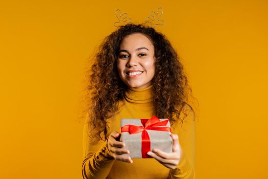 urprised mixed race woman smiling and holding gift box on yellow studio background. Girl with curly hairstyle in sweater. Christmas mood. High quality photo