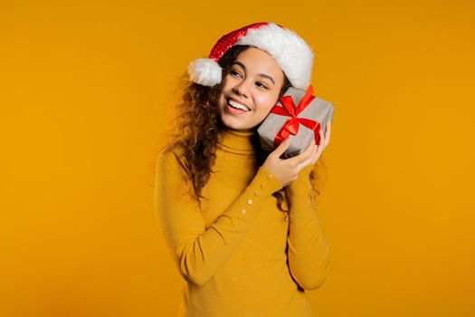 Excited woman in Santa hat received gift box with bow. She is happy and flattered by attention. Girl with present on yellow background. Studio shot. High quality photo