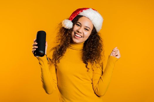 Young cute girl smiling and dancing with wireless portable speaker on yellow studio background. Woman in Santa hat. New year mood. High quality photo