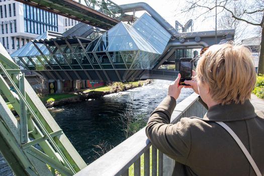 middle-aged blonde woman making a report of the Wuppertal Suspension Railway train in Germany, using mobile phone, The unique electric monorail system is landmark . High quality photo
