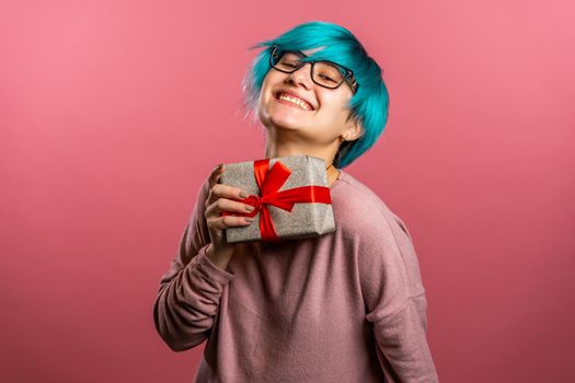 Young unusual woman with blue hair smiling and holding gift box on pink studio background. Girl cozy sweater and glasses. Christmas mood. High quality photo