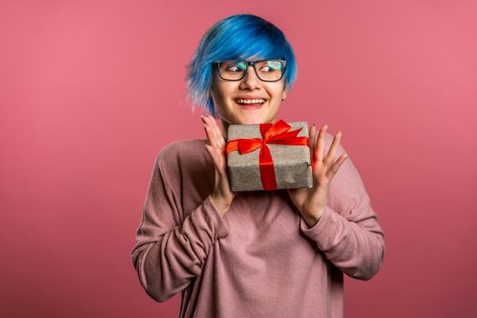 Young unusual woman with blue hair smiling and holding gift box on pink studio background. Girl cozy sweater and glasses. Christmas mood. High quality photo