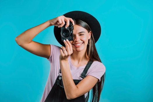 Young pretty woman in overall takes pictures with DSLR camera over blue background in studio. Girl smiling and having fun as photographer. High quality photo