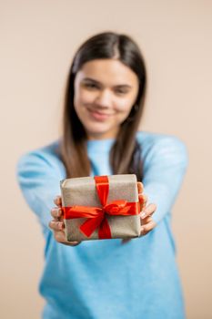 Excited woman holding gift box and gives it by hands to camera on light wall background. Girl smiling, she is happy with present. Studio portrait.