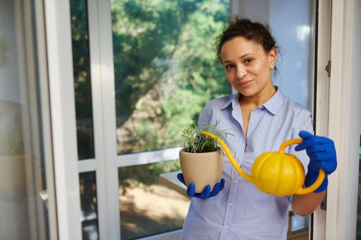 Beautiful woman of African descent in rubber gloves smiles cutely at the camera, poses with rosemary in a pot and a watering can, leans against the door on the veranda of the cottage. Household chores