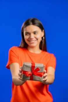 Excited woman holding gift box and gives it by hands to camera on blue wall background. Girl smiling, she is happy with present. Studio portrait.