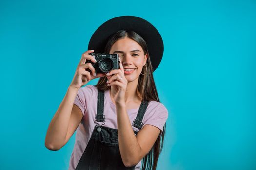 Young pretty woman in overall takes pictures with DSLR camera over blue background in studio. Girl smiling and having fun as photographer. High quality photo