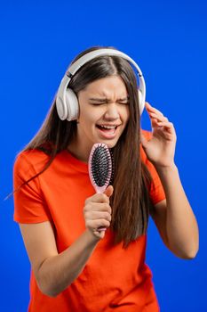European woman singing and dancing with hair brush or comb instead microphone at blue studio background. Lady in headphones having fun, listening to music, dreams of being celebrity. High quality