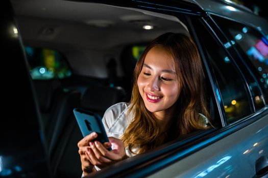 Asian businesswoman working late commuting from office in Taxi backseat with mobile phone in city at night after late work, Happy beautiful woman texting smartphone sitting car back seat in urban