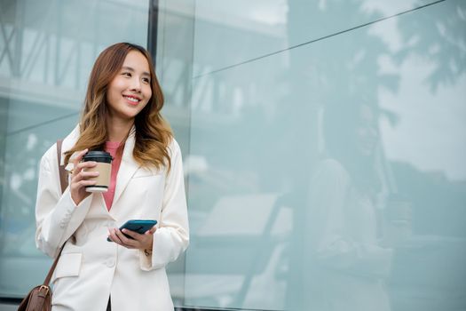 Young business woman smiling holding mobile phone with coffee take away going to work early in morning, Asian businesswoman with smartphone and cup coffee standing against street building near office
