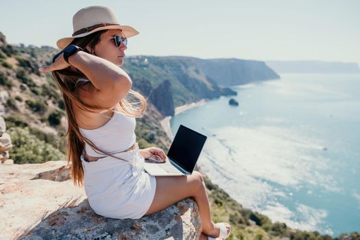 Successful business woman in yellow hat working on laptop by the sea. Pretty lady typing on computer at summer day outdoors. Freelance, travel and holidays concept.