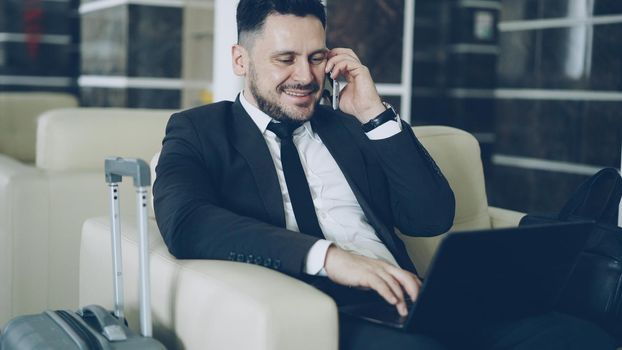 Tilt up of confident attractive businessman talking mobile phone and using laptop computer while sitting on armchair in luxury hotel with luggage suitcase near him. Travel, business and people concept