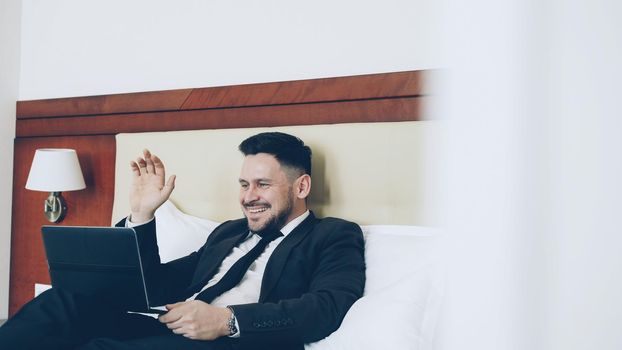 Happy cheerful businessman talking with family via online video chat using laptop computer lying on bed in hotel room. Travel, business and people concept