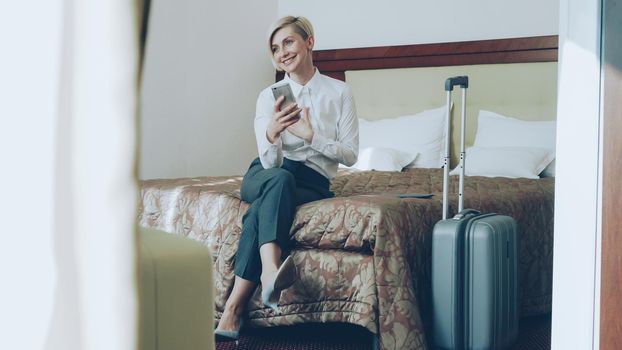 Happy cheerful businesswoman talking with family via online video chat using smrtphone camera sitting on bed in hotel room. Travel, business and people concept