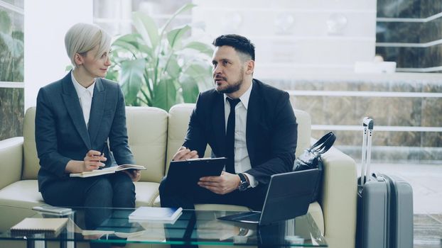 Confident businessman with clipboard talking to female colleague with notepad in hotel lobby prepraing for business meeting they arrived to. Luggage is near couch