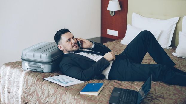 PAN of confident young businessman talking mobile phone while lying on bed in hotel room . Travel, business and people concept