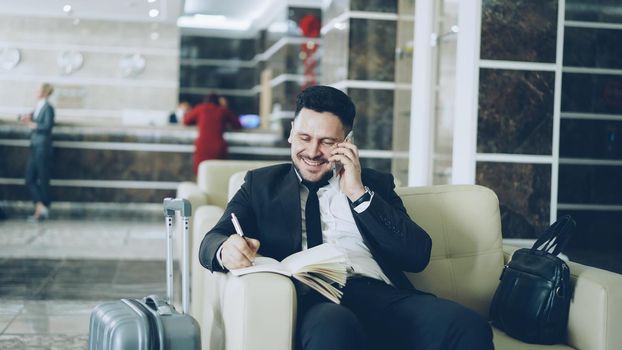 Pan shot of cheerful confident businessman talking mobile phone smiling and writing notes in notepad while sitting on armchair in hotel reception. Travel, business and people concept