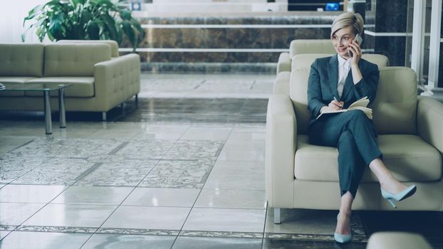 Attractive cheerful businesswoman sitting in armchair in hotel lobby talking on mobile phone and writing in notepad smiling and looking aside. Business, travel and people concept