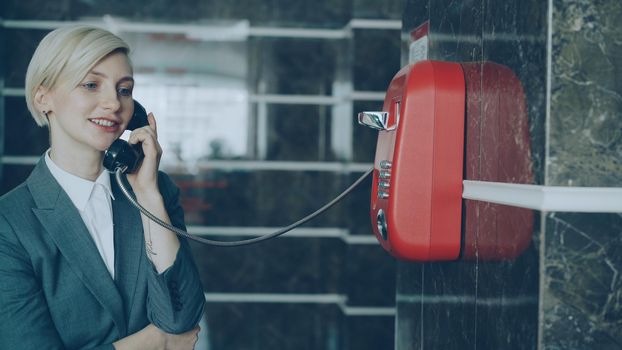 Cheerful blonde businesswoman talking with staff with wired red telaphone at reception in hotel lobby. Business, travel and people concept