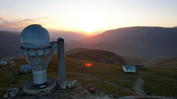 Two large telescope domes at sunset. Drone view of Assy-Turgen Observatory. Beautiful red sunset. Green hills and clouds. Tourists watch the sun. There is a large tent camp and cars nearby. Kazakhstan