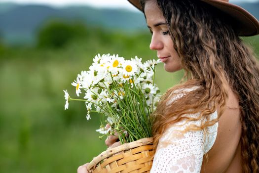 A middle-aged woman in a white dress and brown hat holds a large bouquet of daisies in her hands. Wildflowers for congratulations.