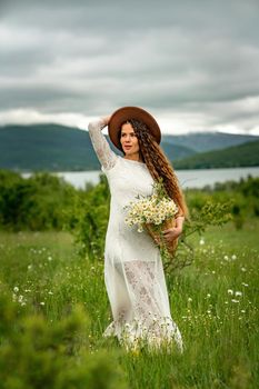 A middle-aged woman in a white dress and brown hat holds a large bouquet of daisies in her hands. Wildflowers for congratulations.