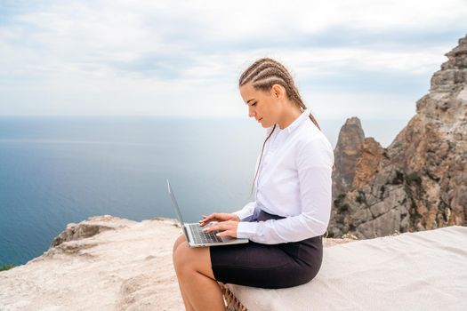 A woman is sitting and typing on a macbook keyboard on a terrace with a beautiful sea view. Wearing a white blouse and black skirt. Freelance travel and vacation concept, digital nomad