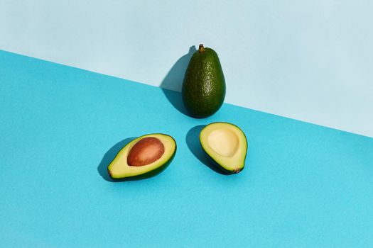 Composition of fresh fruits, whole avocado and two halfs of cutted avocado on blue background. Mock up, two-colored pastel