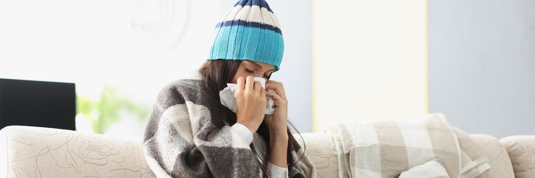 Sick woman in warm hat blowing her nose into paper napkin at home. Treatment of seasonal colds at home concept