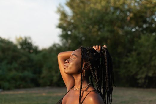 Side view of a young black female holding her braids with her hand in a ponytail with closed eyes in the park at sunset