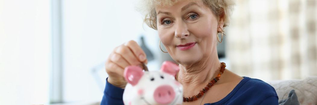 Elderly woman putting coin into piggy bank at home. Saving on retirement concept