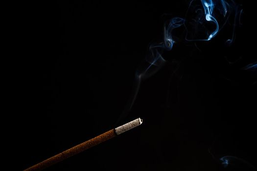 close-up of a burning incense with white smoke coming out illuminated on a black background