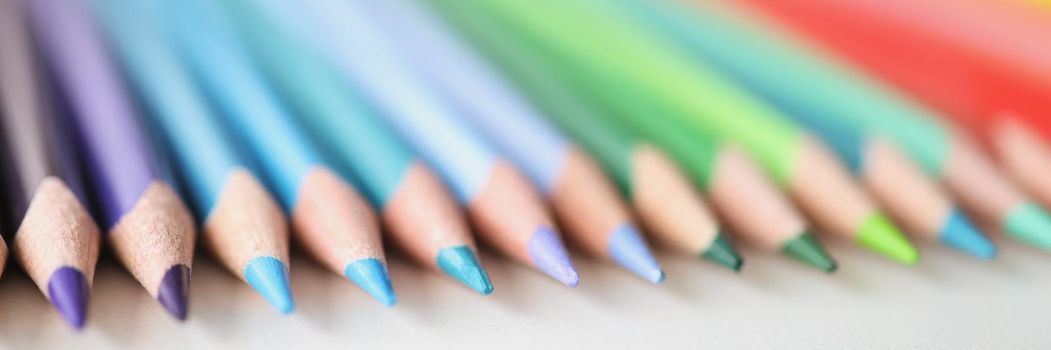 Many sharp multicolored pencils lying over colors of rainbow closeup background. School of fine arts concept