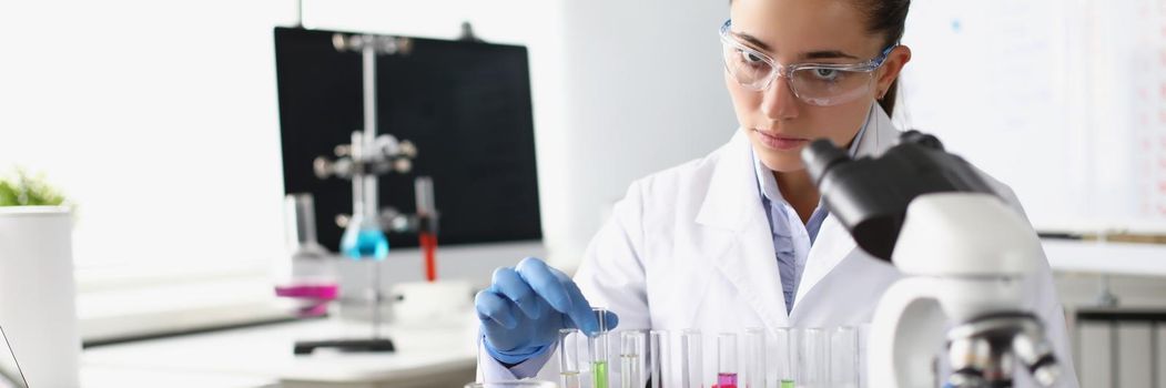 Chemist woman holding test tube with liquid in front of microscope in laboratory. Pharmaceutical industry concept