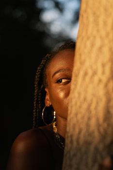 Close-up portrait of a young black woman hiding half of her face behind a tree while looking to the side in the park