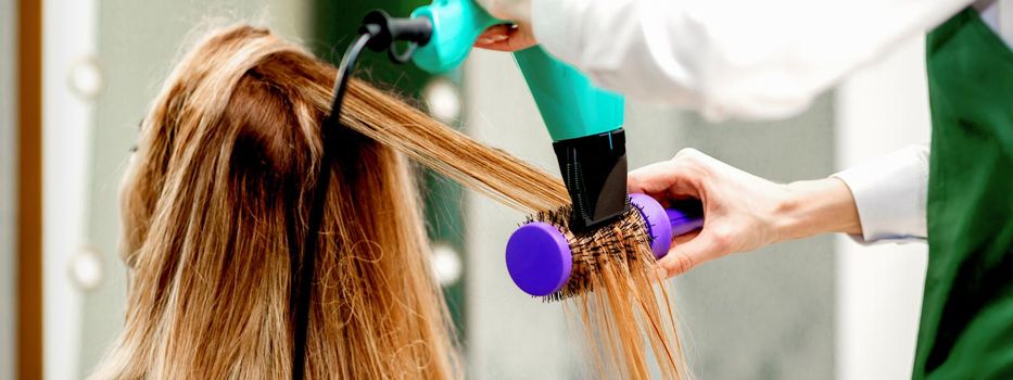 Back view of young woman receiving drying hair with a hairdryer and hairbrush in a hair salon