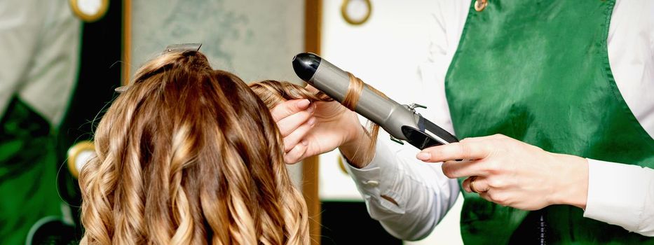 Back view of female hairdresser's hands curling women's hair with curling iron in a hair salon