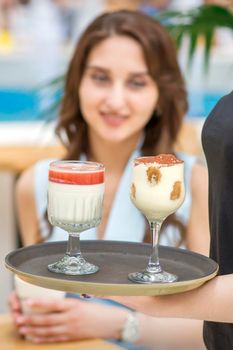 A young waitress carrying a tray with two different desserts in a two glass on the background of a young female client